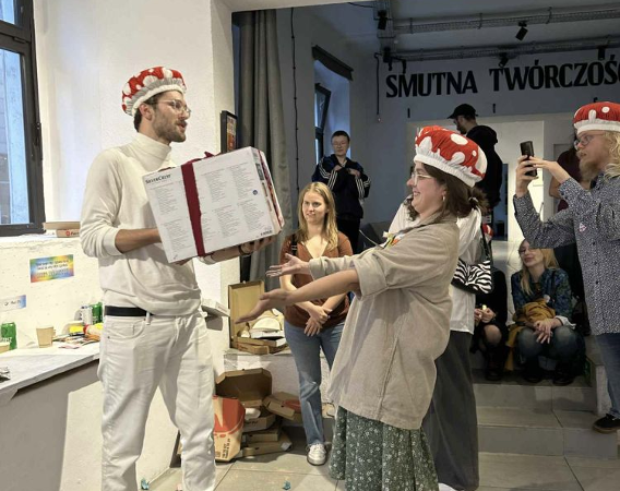 Młoda kobieta wręcza prezent młodemu mężczyźnie. Oboje mają na głowach czapki w kształcie muchomorów.