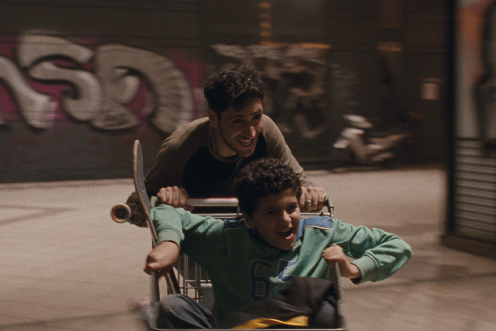 Młody mężczyzna i jeszcze młodszy chłopak bawią się wózkiem sklepowym, jeden wiezie drugiego i nabierają prędkości.