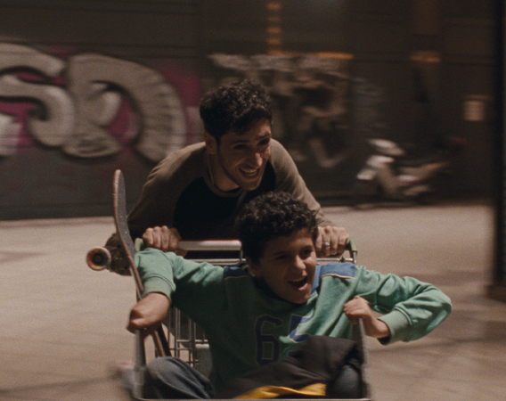 Młody mężczyzna i jeszcze młodszy chłopak bawią się wózkiem sklepowym, jeden wiezie drugiego i nabierają prędkości.