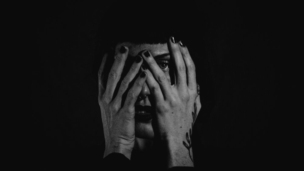Artystka zakrywa twarz rękami (na jednej z nich roślinny tatuaż) z pomalowanymi na czarno paznokciami, widać tylko jej prawe oko i fragment ust. Zdjęcie czarne, ponure, tajemnicze.