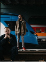 Mrokas stoi na peronie i Mrokas siedzi na peronie. W tle pociągi.