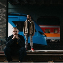 Mrokas stoi na peronie i Mrokas siedzi na peronie. W tle pociągi.