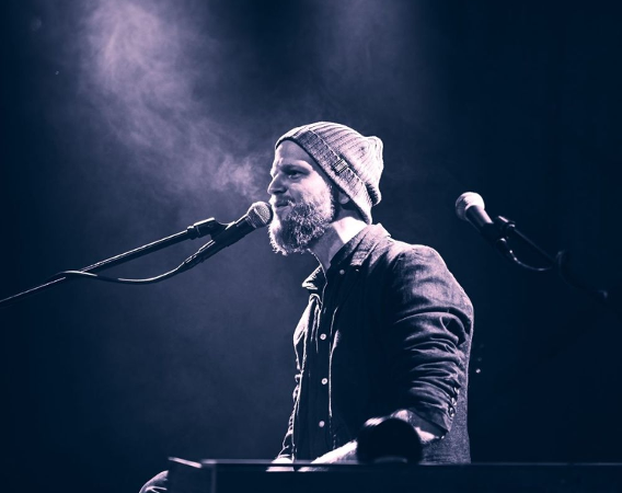 Muzyk ma rudą brodę, na głowie czapkę. Stoi na scenie, śpiewając i jednocześnie grając na klawiszach.