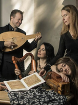 Na zdjęciu grupa muzyków. Znajdują się wokół stołu. Ktoś gra w zamyśleniu - a ktoś patrzy w dal.