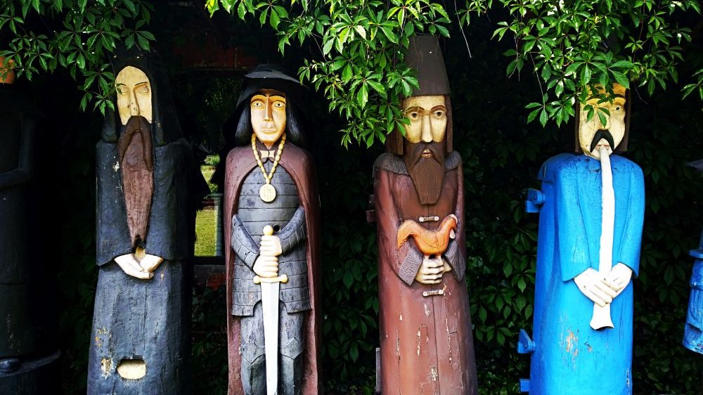 Na fotografii znajdują się cztery kolorowe, drewniane, starodawne ule figuralne przedstawiające mężczyzn. Pierwszy od lewej ubrany jest w czarną szatę mnicha, drugi w zbroję oraz brązową pelerynę, trzyma miecz.. - grafika artykułu