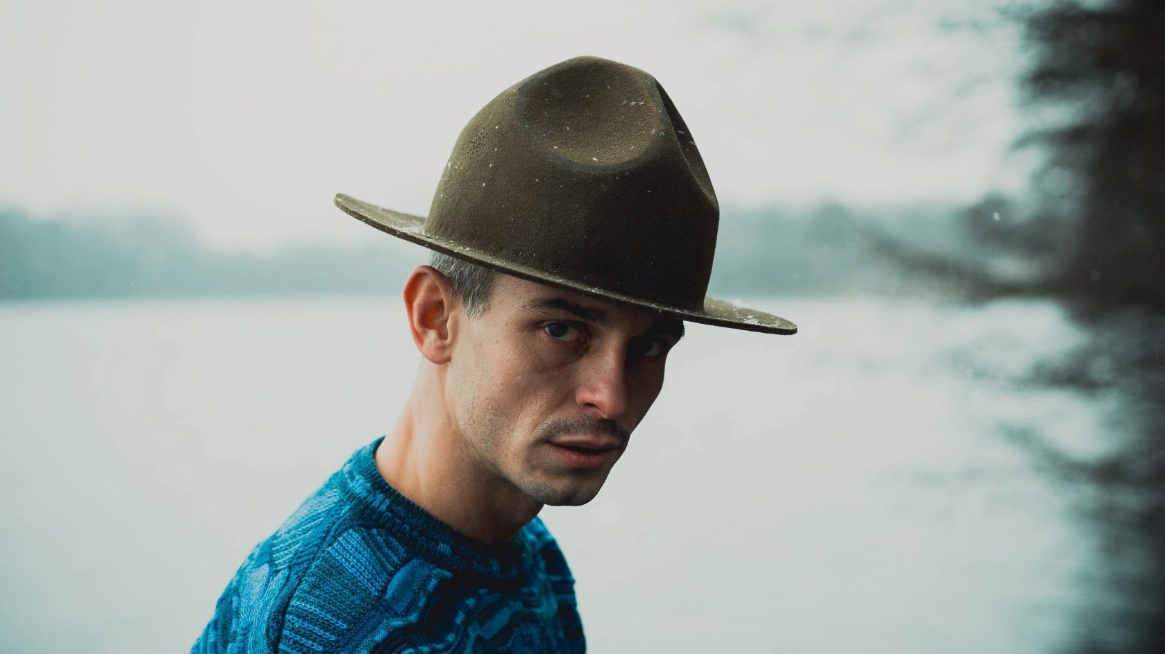 Mężczyzna w kapeluszu patrzy w obiektyw. W tle jezioro, pogoda pochmurna.