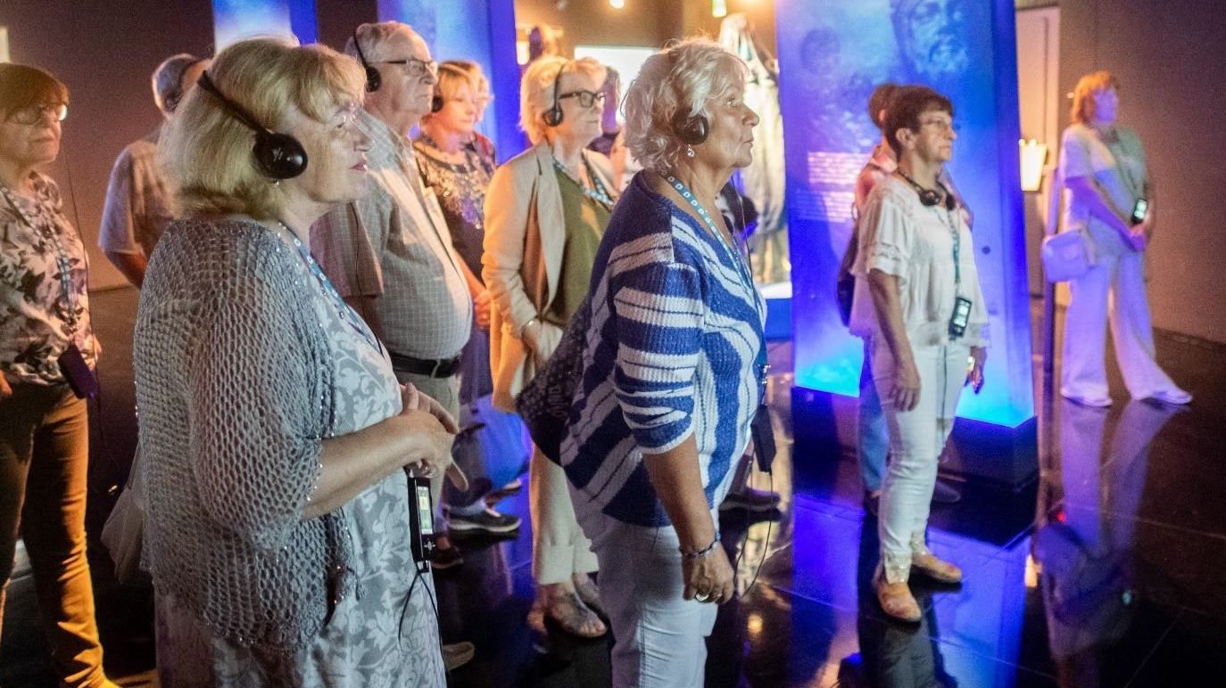 Grupa seniorów zwiedzających ekspozycję, wpatrzonych w jeden punkt, na głowach mają słuchawki, a na szyi zawieszone audioprzewodniki.