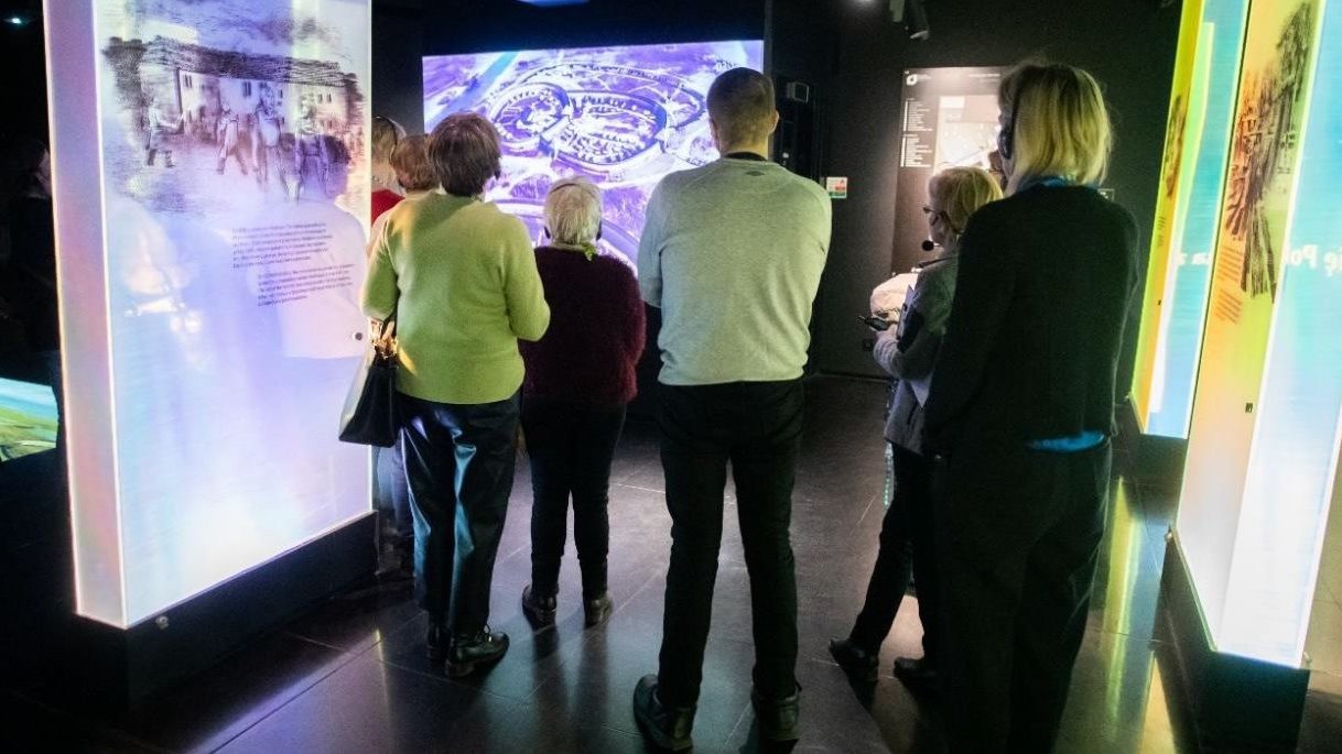 Grupa zwiedzających uchwyconych przez fotografa od tyłu zwiedza nowoczesną, multimedialną ekspozycję, przed nimi ekran z obrazem ogrodu na Ostrowie Tumskim z czasów Mieszka I