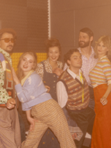 Grupa aktorów i aktorek przebranych w kolorowe stroje w stylu lat 80.