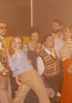 Grupa aktorów i aktorek przebranych w kolorowe stroje w stylu lat 80.