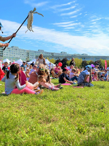 W osiedlowym parku na trawie siedzą dzieci z rodzicami. Wokół nich poruszają się aktorzy teatralni, którzy odgrywają spektakl z użyciem lalek. W tle długi blok mieszkalny i niebo pokryte delikatnymi chmurkami.