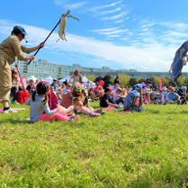 W osiedlowym parku na trawie siedzą dzieci z rodzicami. Wokół nich poruszają się aktorzy teatralni, którzy odgrywają spektakl z użyciem lalek. W tle długi blok mieszkalny i niebo pokryte delikatnymi chmurkami.