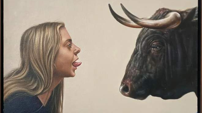 Na obrazie młoda blondynka z profilu, nachyla się i pokazuje język stojącemu naprzeciw niej czarnemu bykowi.