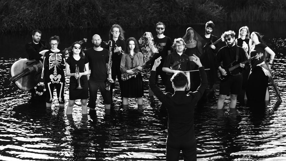 Czarno-biała fotografia, kilkunastu członków orkiestry stoi w wodzie trzymając w rękach instrumenty muzyczne. Na pierwszym planie dyrygent z uniesionymi rękoma. - grafika artykułu