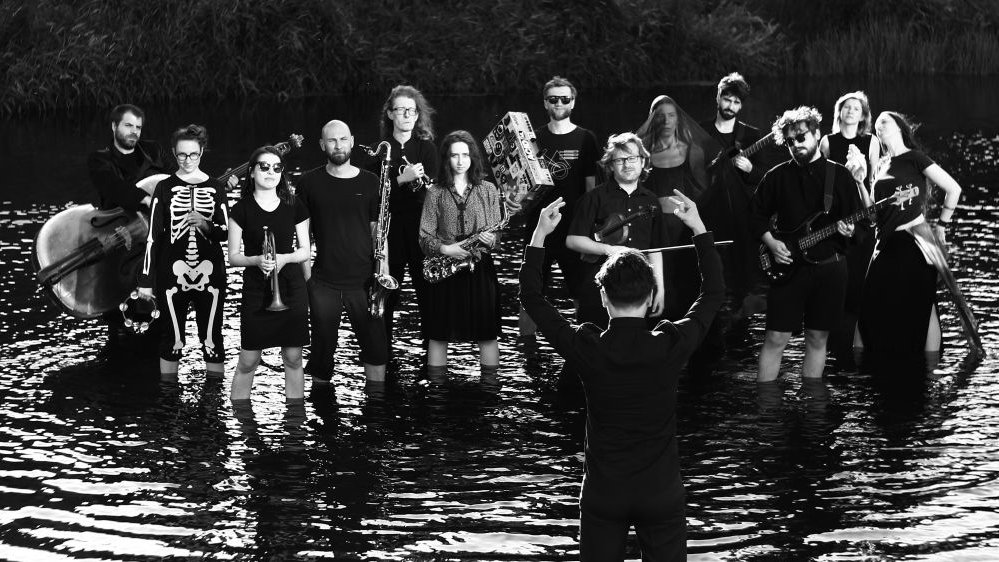 Czarno-biała fotografia, kilkunastu członków orkiestry stoi w wodzie trzymając w rękach instrumenty muzyczne. Na pierwszym planie dyrygent z uniesionymi rękoma.