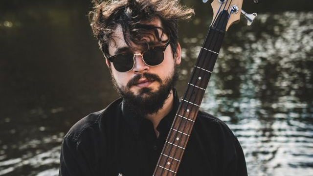 Mężczyzna w okularach przeciwsłonecznych i czarnym stroju stoi w wodzie trzymając w dłoniach gitarę basową.
