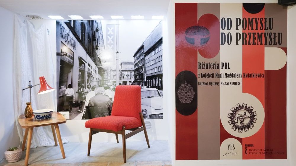 Po prawej plakat reklamujący wystawę, po lewej stolik i fotel z okresu PRL na tle czarno-białej fotografii ulicy dużego miasta.