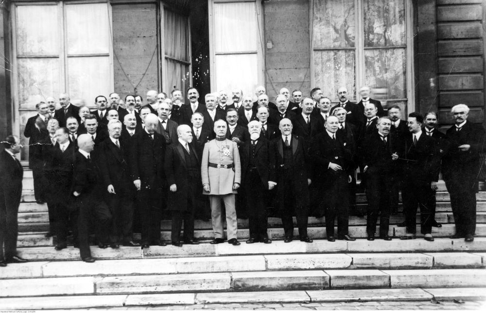 Czarno-białe zdjęcie grupowe. Mężczyźni w czarnych frakach i jeden w jasnym mundurze (w centrum tej grupy) stoją na schodach budyku. - grafika artykułu