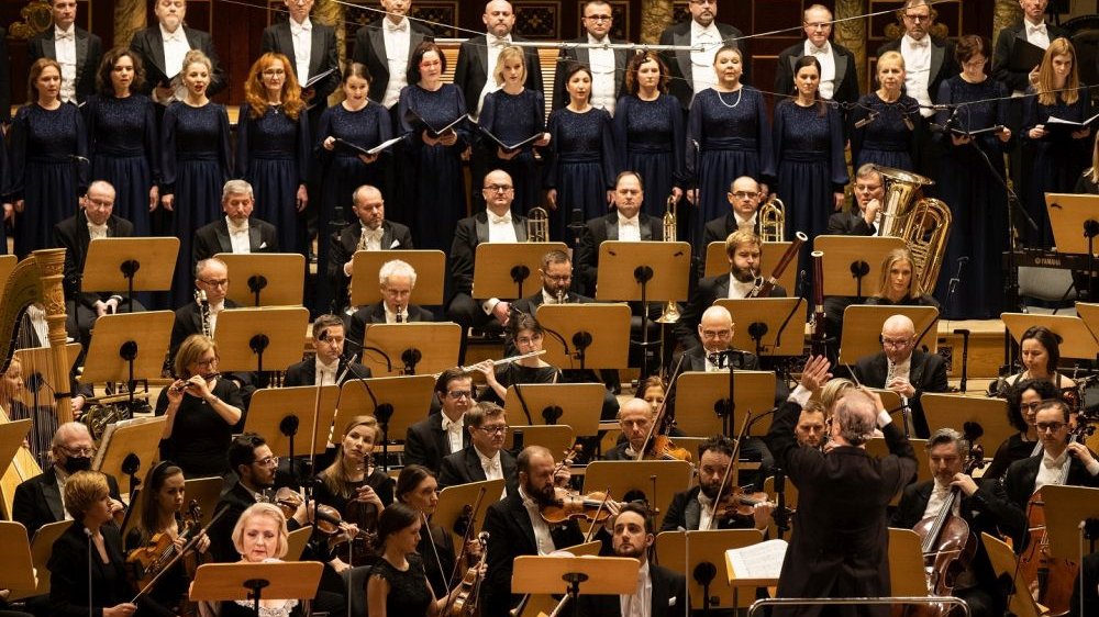 U góry zdjęcia w dwóch rzędach stoją chórzyści i chórzystyki. Chórzyści mają na sobie czarne fraki i białe koszule, chórzystki są z kolei ubrane w granatowe suknie. Pod nimi orkiestra Teatru Wielkiego w eleganckich, ciemnych ubraniach. W rogu zdjęcia - dyrygent.