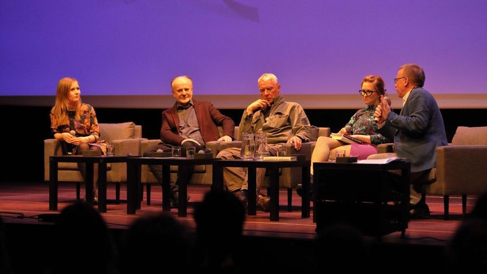 Na scenie siedzi czwórka gości spotkania, dwie poetki oraz dwóch poetów, z boku widać prowadzącego spotkanie profesora Piotra Śliwińskiego w granatowej marynarce.