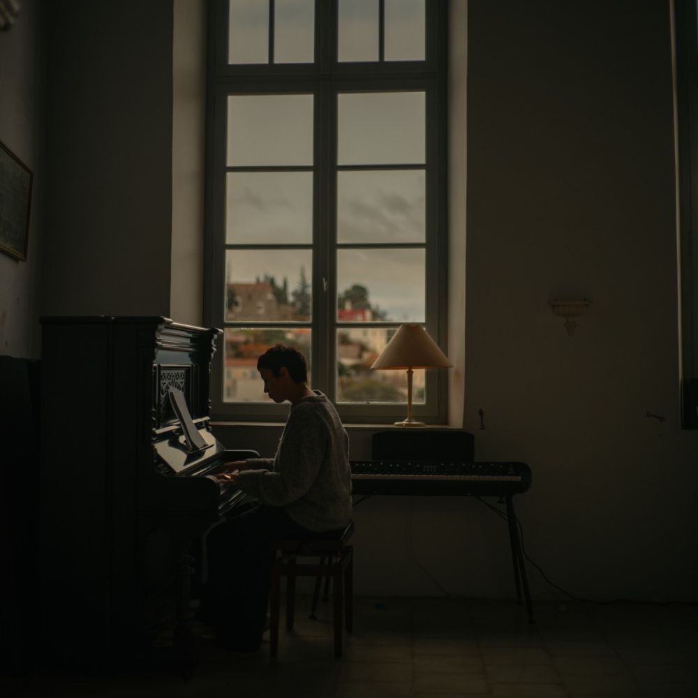 Artystka gra na fortepianie. Pomieszczenie, w którym się znajduje jest ciemne i ponure, przez duże okno widać kilka domów, dzień jest pochmurny. - grafika artykułu