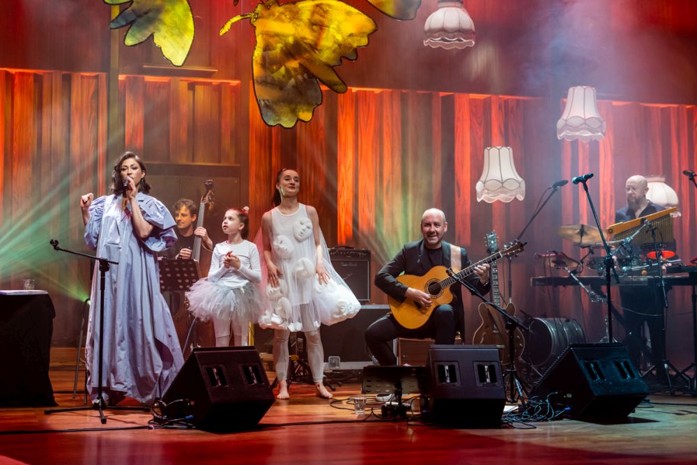 Artystka w długiej, fioletowej sukni, śpiewa obok dwóch dziewczynek ubranych w baletowe sukienki. Na scenie siedzi także gitarzysta. Wszystko jest oświetlone pomarańczowym światłem. - grafika artykułu