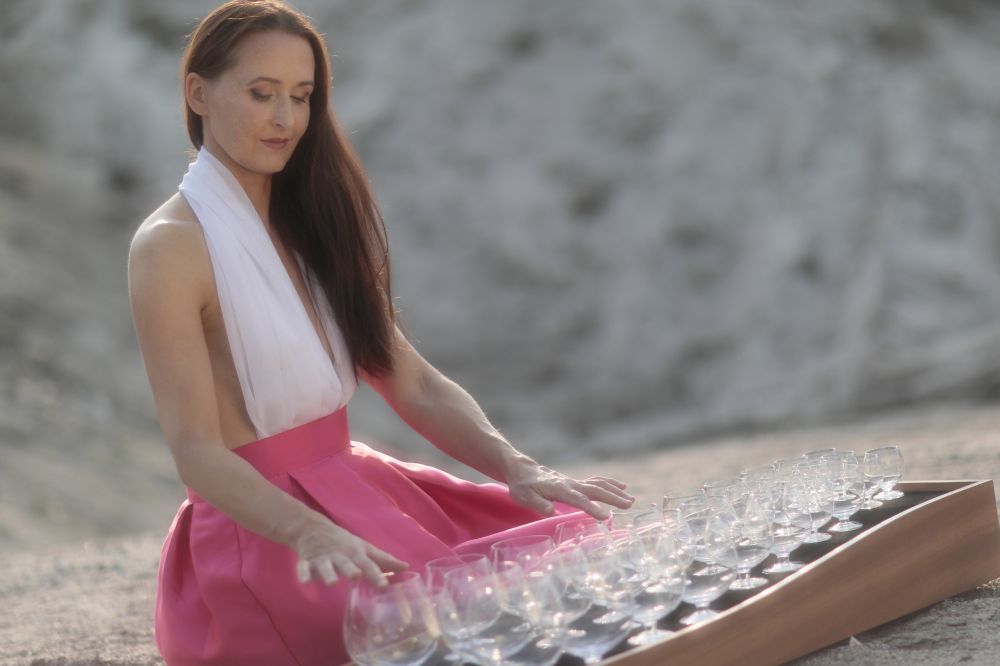 Długowłosa szatynka w biało-różowej sukni siedzi przed platformą z kieliszkami i dotyka ich delikatnie. Zdjęcie słoneczne, pogodne, delikatne. - grafika artykułu