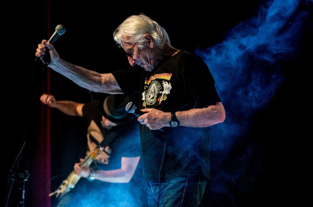 Koncert, na scenie stoi wokalista, trzymający dwa mikrofony, a obok niego gitarzysta. Nad sceną unosi się dym. - grafika artykułu