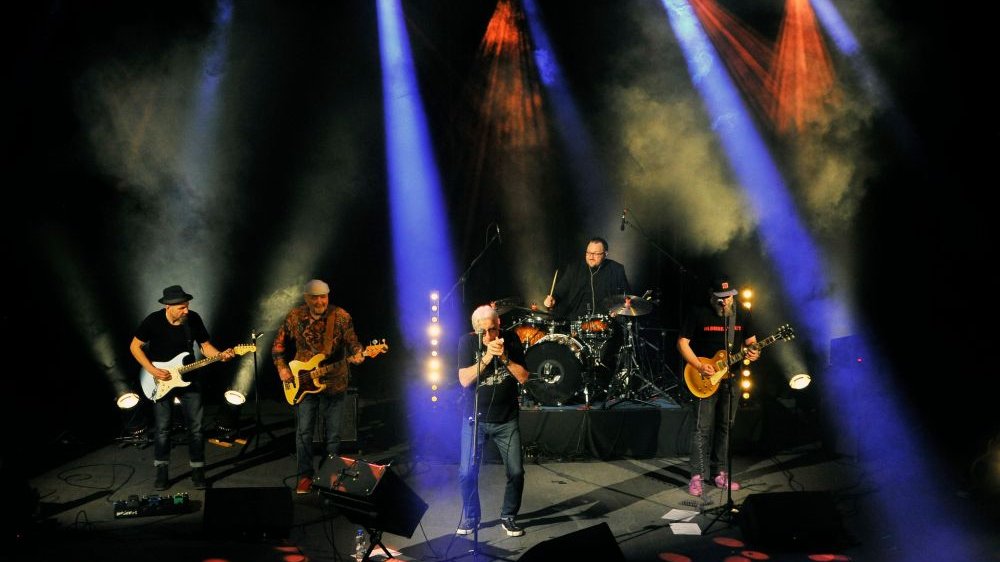 Koncert, na oświetlonej na niebiesko i czerwono scenie pięciu artystów, troje gitarzystów, perkusista oraz wokalista.