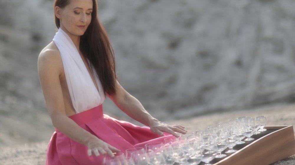 Długowłosa szatynka w biało-różowej sukni siedzi przed platformą z kieliszkami i dotyka ich delikatnie. Zdjęcie słoneczne, pogodne, delikatne. - grafika artykułu