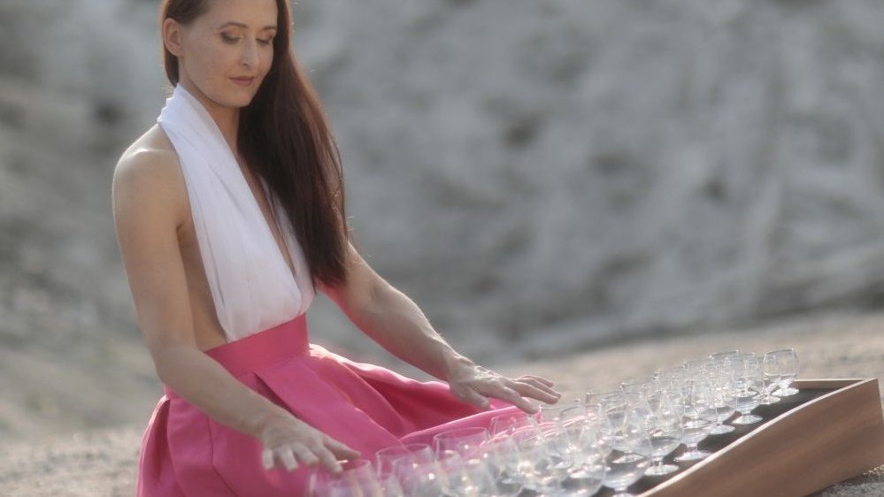 Długowłosa szatynka w biało-różowej sukni siedzi przed platformą z kieliszkami i dotyka ich delikatnie. Zdjęcie słoneczne, pogodne, delikatne.
