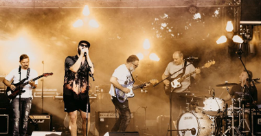 Artyści na scenie, trójka gitarzystów ubranych w białe koszulki oraz wokalista w czapce z daszkiem, ciemnych okularach i hawajskiej koszuli.
