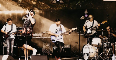 Artyści na scenie, trójka gitarzystów ubranych w białe koszulki oraz wokalista w czapce z daszkiem, ciemnych okularach i hawajskiej koszuli.
