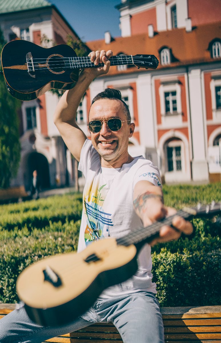 Rozmówca w białej koszulce na dziedzińcu Urzędu Miasta Poznania, trzyma w rękach dwa instrumenty ukulele, przypominające małe gitary - grafika artykułu