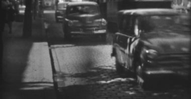 Biało-czarne i poruszone kadry z filmu przedstawiające jadące brukowaną miejską ulicą samochody.