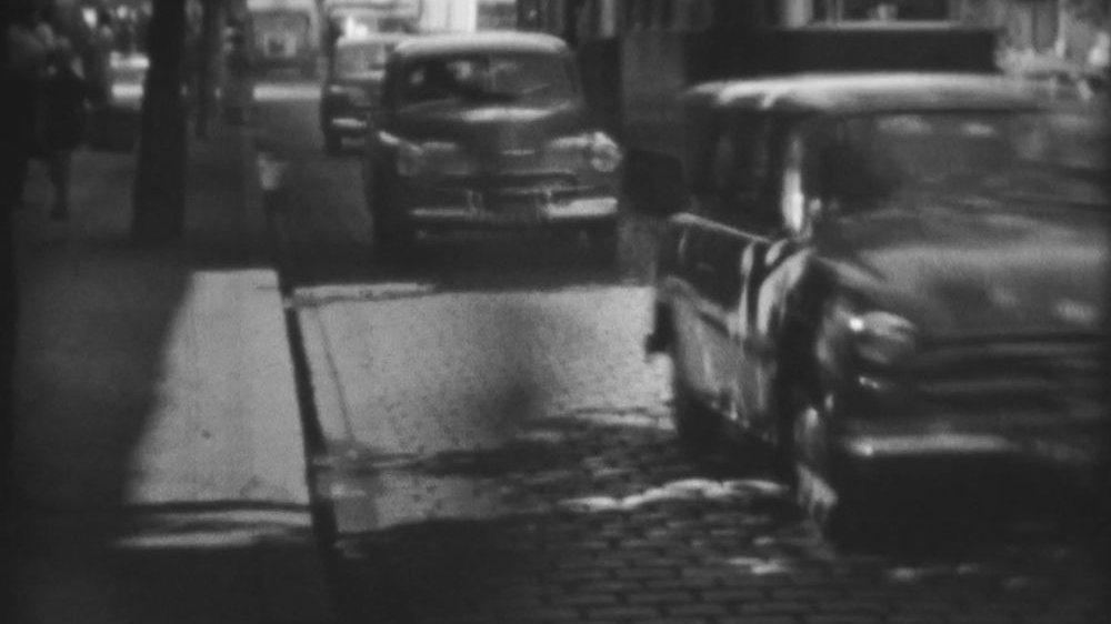 Biało-czarne i poruszone kadry z filmu przedstawiające jadące brukowaną miejską ulicą samochody.