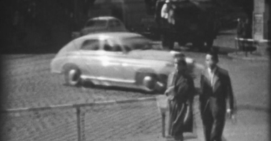 Poruszony, biało-czarny kadr z filmu 8-milimetrowego. Para idąca chodnikiem w pobliżu skrzyżowania, z tyłu jadące samochody.