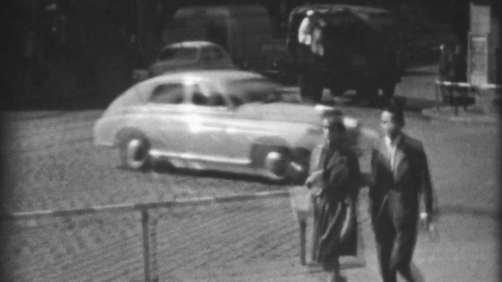 Poruszony, biało-czarny kadr z filmu 8-milimetrowego. Para idąca chodnikiem w pobliżu skrzyżowania, z tyłu jadące samochody.