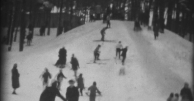 Niewyraźne zdjęcie będące fragmentem klatki filmowej. Na nim ludzie zjeżdżający na sankach i nartach po śniegu z górki położonej w mieście.
