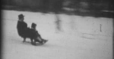 Niewyraźne zdjęcie będące fragmentem klatki filmowej. Na nim ojciec z dzieckiem zjeżdżający na sankach po śniegu z górki położonej w mieście.