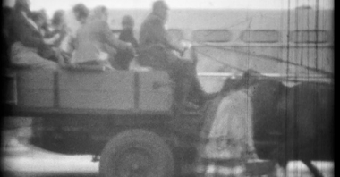 Niewyraźny, czarno-biały kadr ze starego filmu. Wóz zaprzężony w dwa konie wiezie ludzi, mężczyznę w czapce, kobiety w chustach na głowie i dziecko. Obok wozu idą pieszo dwie kobiety.