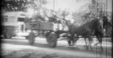 Poruszony, czarno-biały kadr ze starego filmu. Ludzie jadą wozem ciągniętym przez dwa konie. Z tyłu drzewa i autobus.