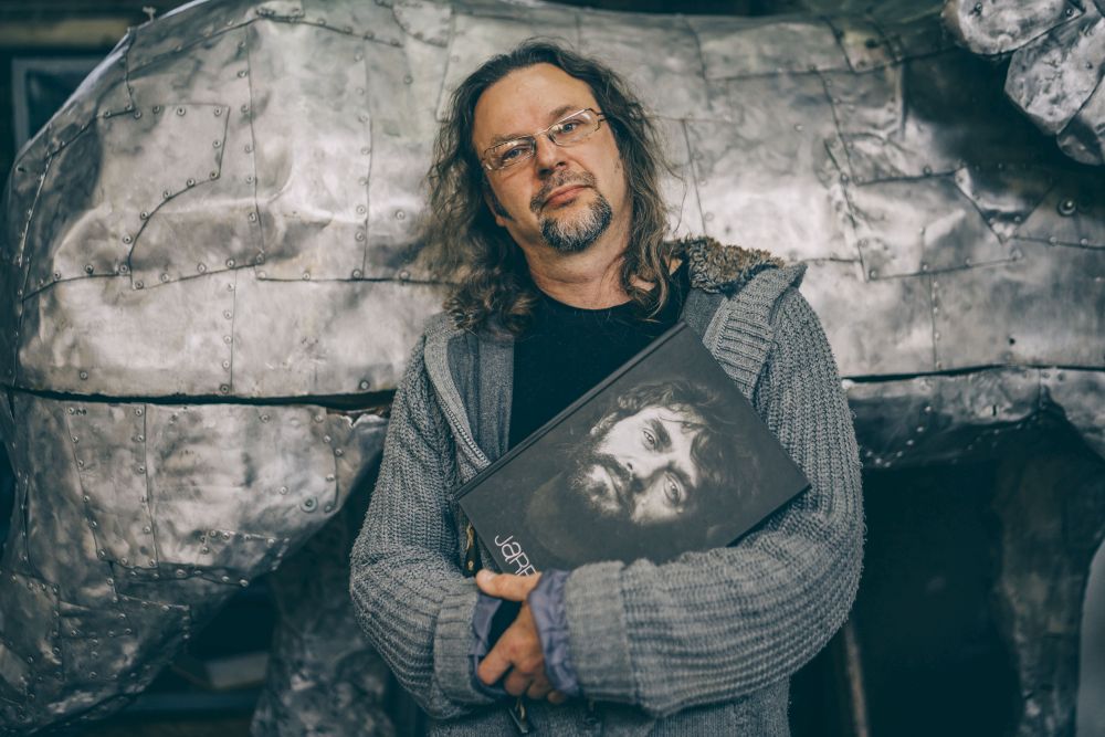 Mężczyzna w długich włosach i z brodą opiera się o metalową konstrukcję. W rękach trzyma książkę z portretem Jarosława Maszewskiego na okładce. - grafika artykułu