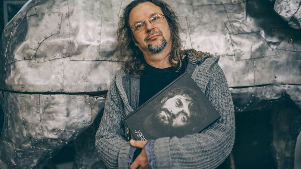 Mężczyzna w długich włosach i z brodą opiera się o metalową konstrukcję. W rękach trzyma książkę z portretem Jarosława Maszewskiego na okładce.