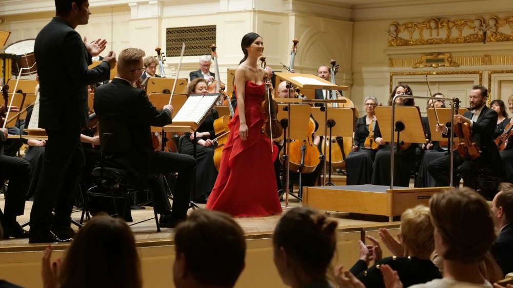 Skrzypaczka stoi wyprostowana przed oklaskującą ją publicznością. Otacza ją orkiestra, niedaleko niej stoi dyrygent, także klaszcze.