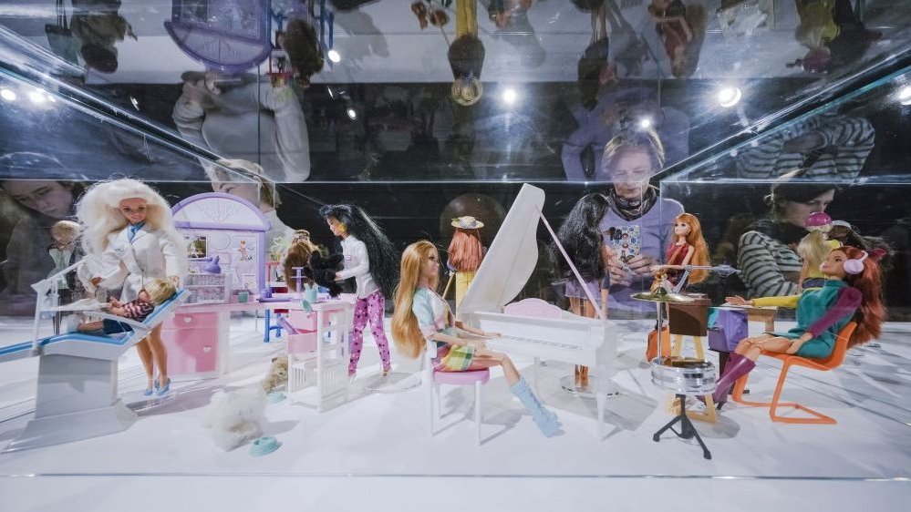 Wiele lalek Barbie na wystawie, pod szklaną gablotą. Z tyłu, za gablotą stojące kobiety zwiedzające wystawę. Lalki są bardzo kolorowe, mają atrybuty, na przykład fortepian czy fotel dentystyczny.