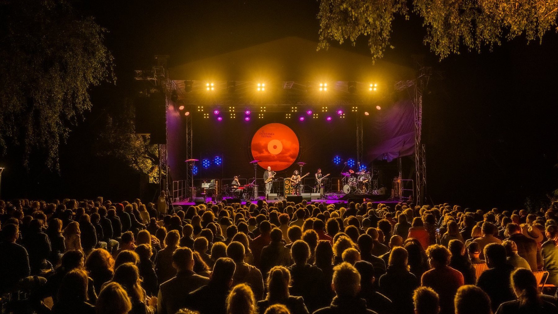 Liczna publiczność festiwalu Enter Enea siedząca na krzesłach zwrócona w stronę oświetlonej mocnymi reflektorami sceny, na której widać kilku muzyków, w tym saksofonistę, perkusistę i dwójkę gitarzystów.