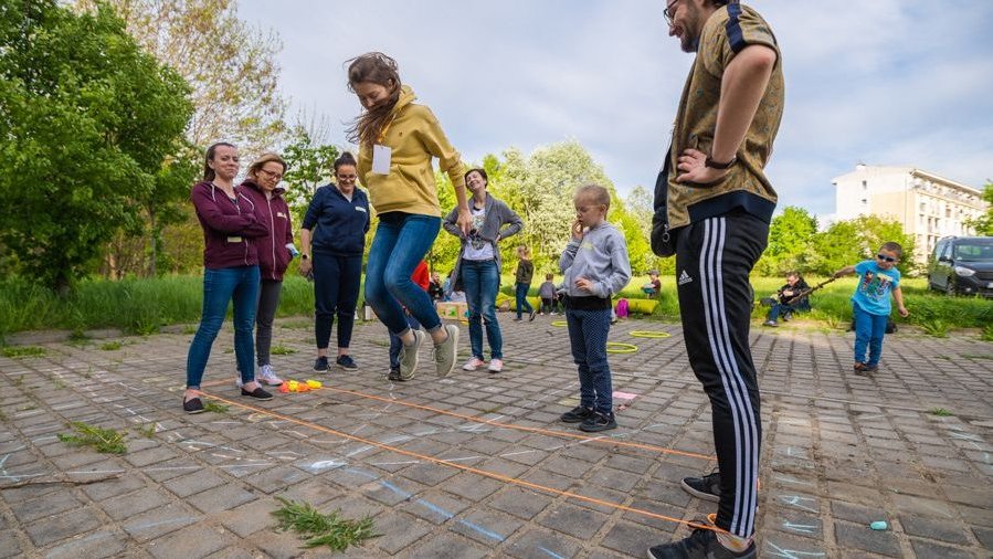 Słoneczny dzień w parku, dzieci oraz animatorzy grają w narysowaną kredą na chodniku grę. Jedna z dziewczynek wysoko podskakuje.