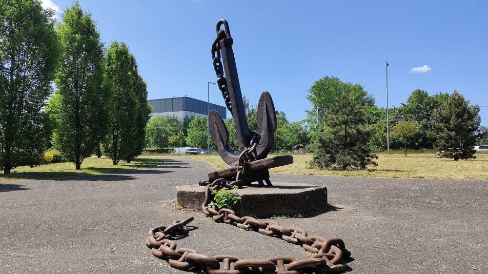 Na środku, na małym cokole, duża metalowa kotwica statku z przyłączonym do niej grubym łańcuchem. Z tyłu trawa i drzewa. Błękitne niebo, dużo słońca.