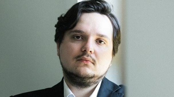 Mężczyzna w garniturze i białej, rozpiętej pod szyją koszuli patrzy prosto w obiektyw.